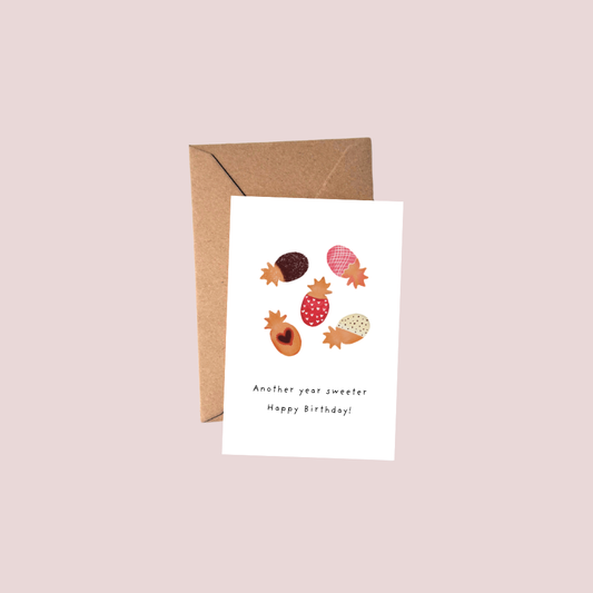 A white greeting card with a brown kraft envelope, featuring an illustration of various acorns and small acorn-shaped ornaments with the text 'Another year sweeter Happy Birthday!' on a light pink background.