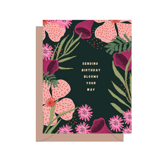 A floral birthday card with a dark green background, pink and magenta flowers, green leaves, and gold text reading 'SENDING BIRTHDAY BLOOMS YOUR WAY,' shown with a kraft envelope behind it on a white background.