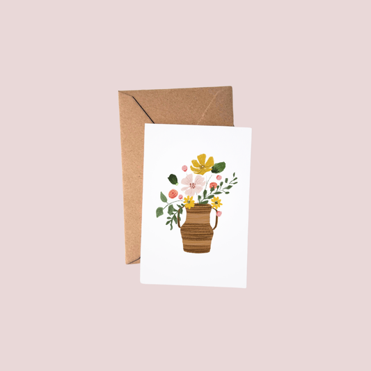 A white greeting card featuring a watercolor illustration of a brown striped vase filled with yellow, pink, and white flowers and green leaves, shown with a brown kraft envelope on a light pink background.