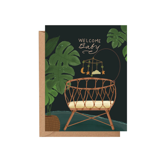 Welcome Baby greeting card featuring a wicker bassinet, nursery mobile, and tropical monstera leaves on a dark green background.